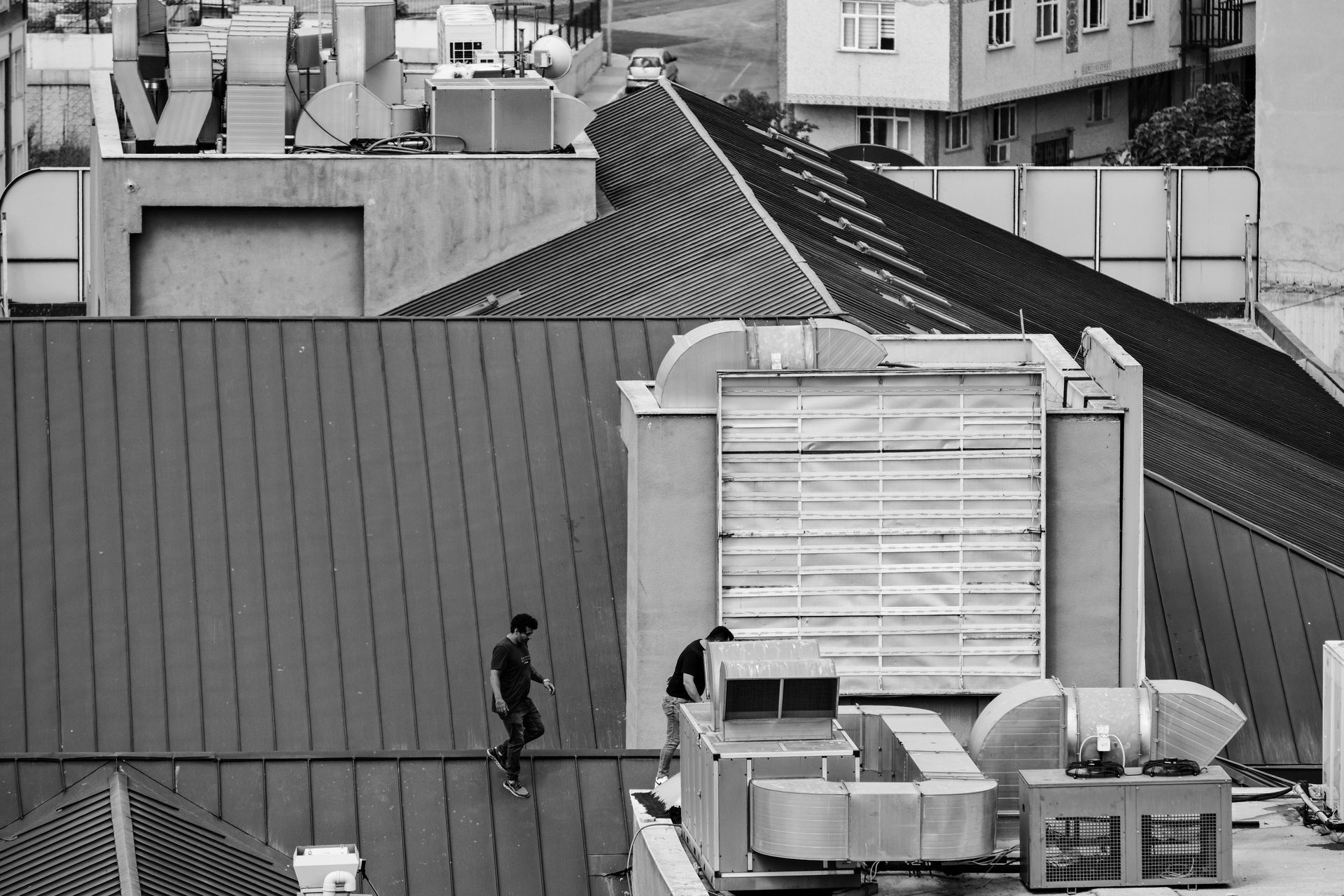 Two people walk across a metal roof next to large ventilation equipment.
