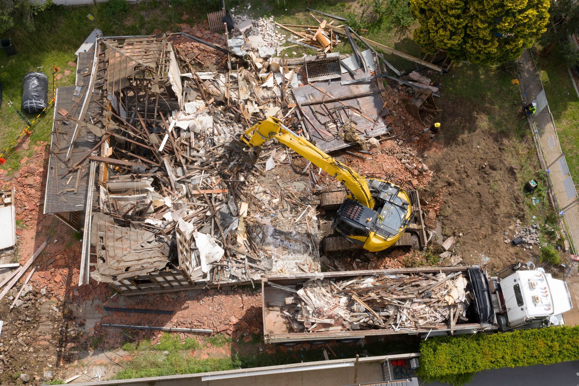 Demolished house with an excavator loading debris into a truck. Yellow machinery, brick foundation, green hedge.