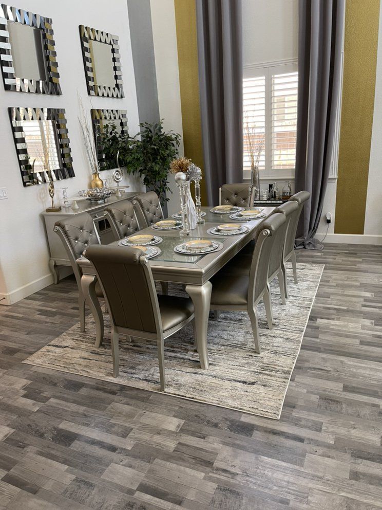 Elegant dining room: silver table, upholstered chairs, patterned rug, mirrored wall decor, gray curtains.