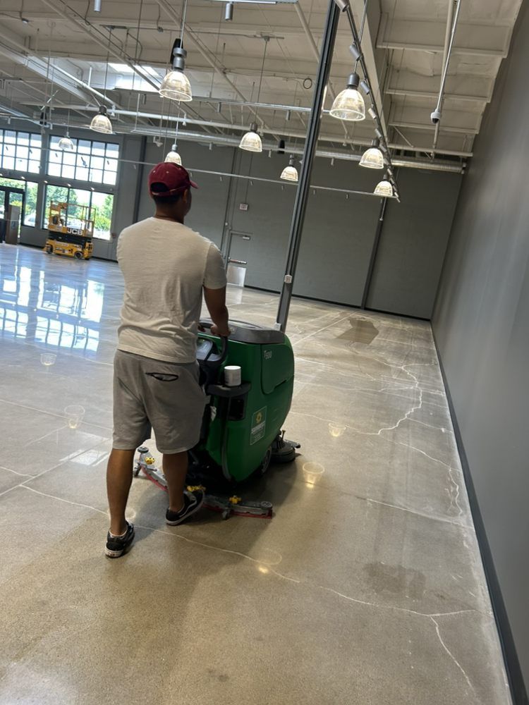 Person operating a floor cleaning machine in a large, empty room with concrete flooring and overhead lights.