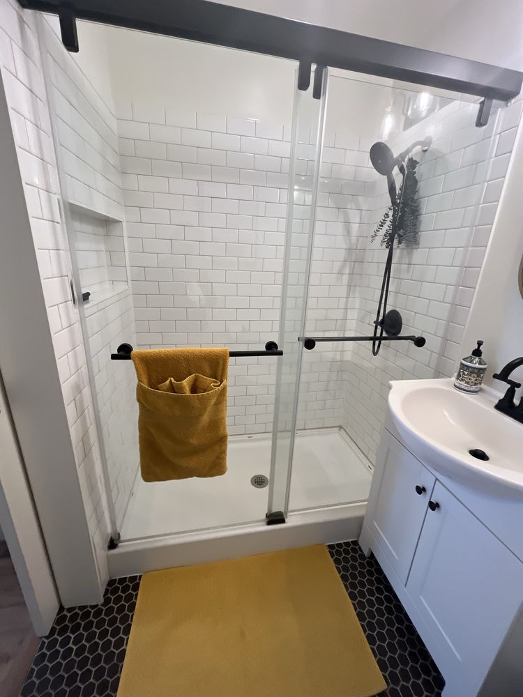 Bathroom with a glass shower door and white tile. Yellow rug and towel, black fixtures.