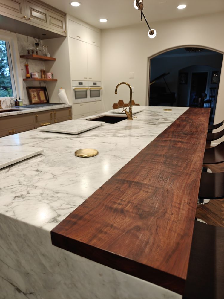 Kitchen island with white marble countertop and a wooden extension with seating. Gold faucet, sink.