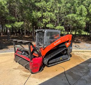 Orange skid steer with brush cutter attachment parked on a sunlit paved area beside trees.