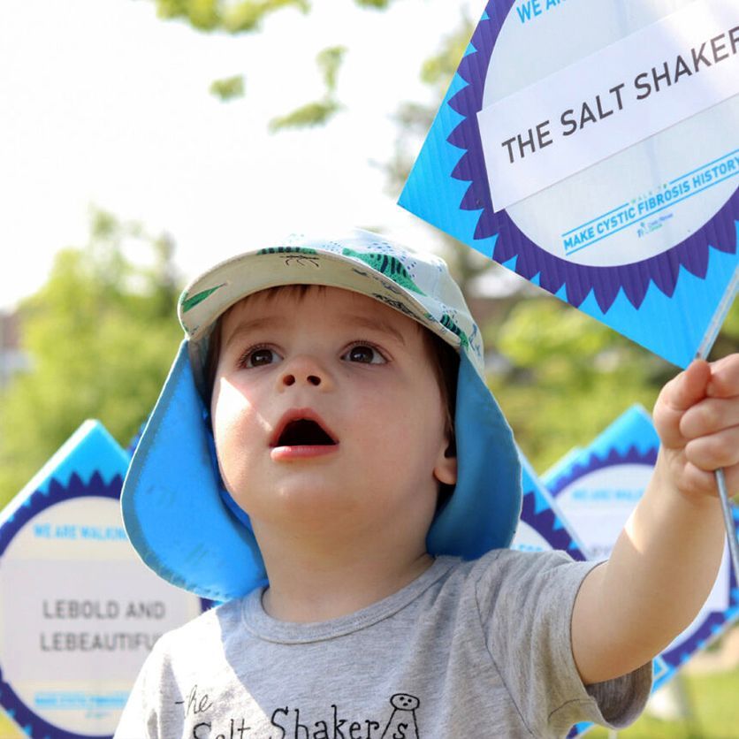 A toddler wearing his Walk gear holding his team sign in the air.