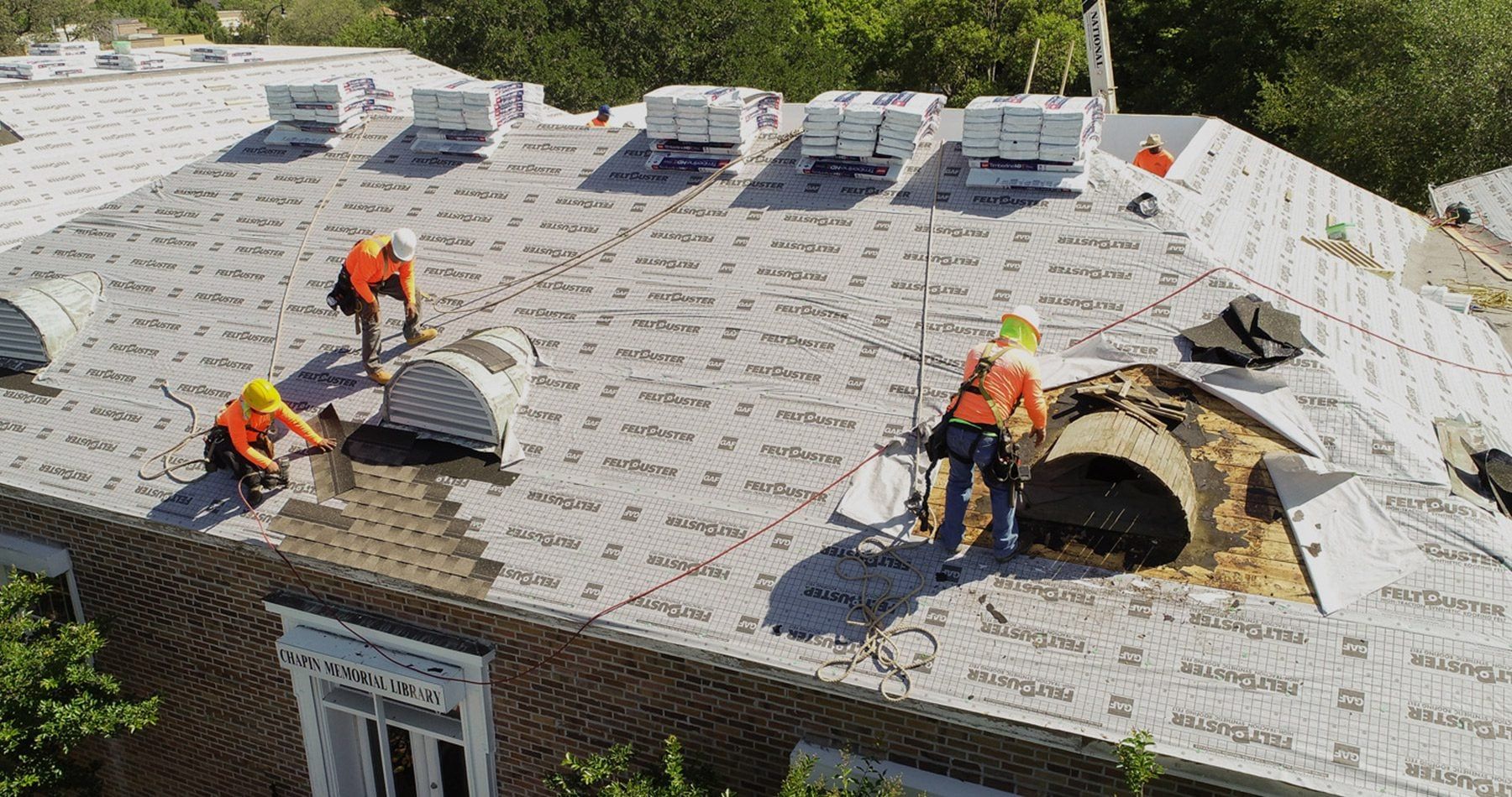 Roofers in orange vests and hard hats working on a roof, surrounded by supplies.