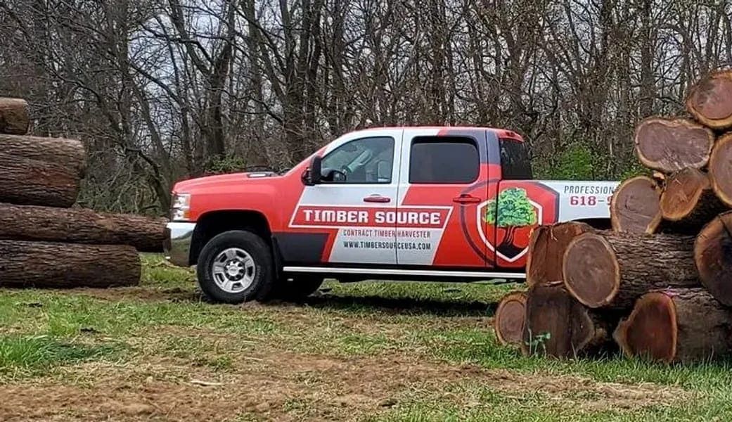 Red and black Timber Source truck parked next to logs in a grassy area, trees in the background.