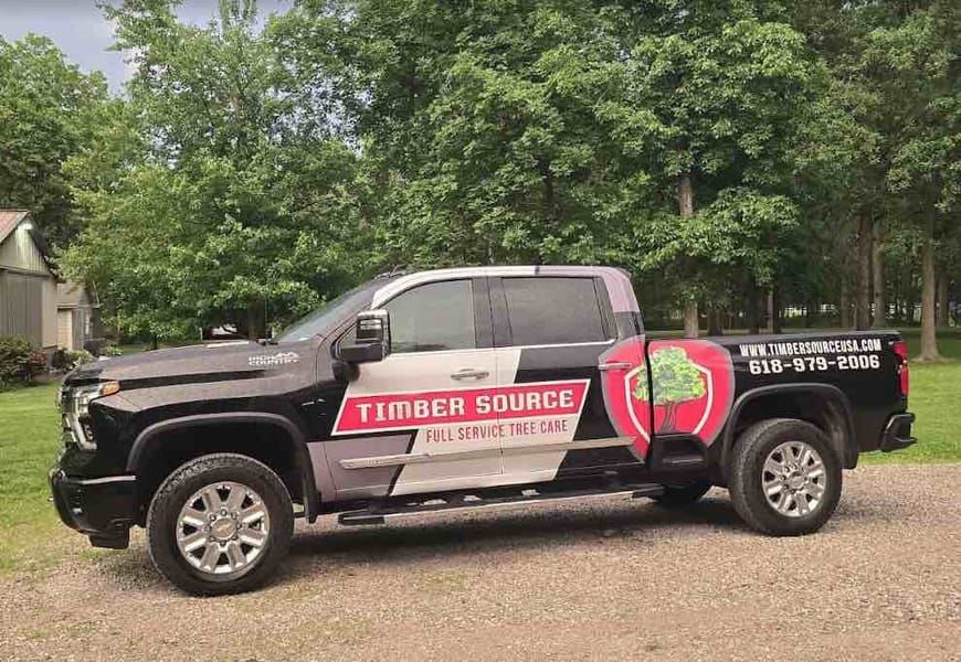 Red and white pickup truck with company logo parked near stacks of logs.