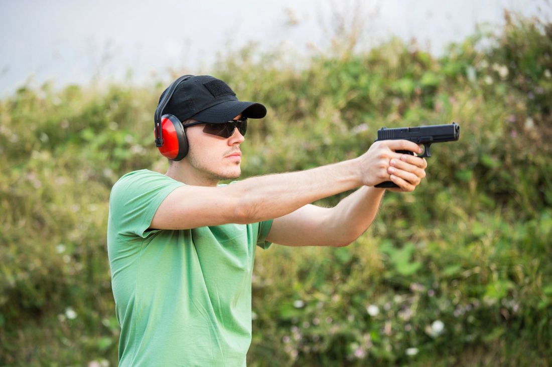 Uomo con camicia verde e berretto nero che punta una pistola all'aperto, con indosso protezioni acustiche rosse.
