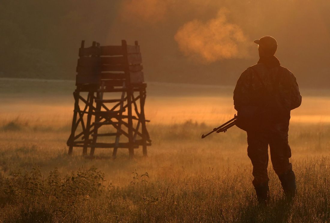 Sagoma armata in un campo nebbioso al tramonto, vicino a una torre di guardia in legno con del fumo sullo sfondo.