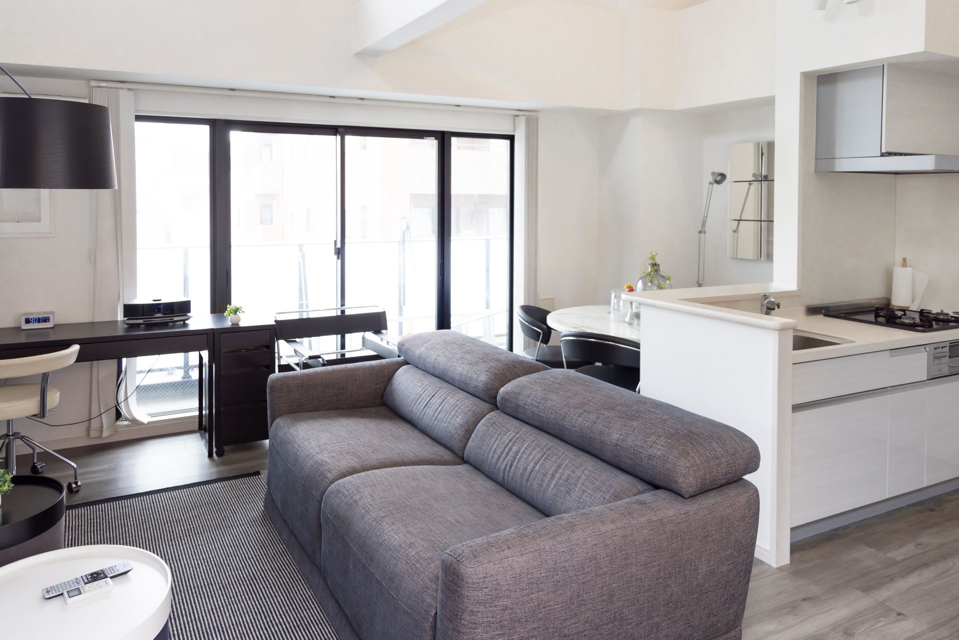 A modern, minimalist living area with a grey sofa, black desk, white coffee table, and an adjacent kitchen. A modern, minimalist living area with a grey sofa, black desk, white coffee table, and an adjacent kitchen.