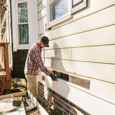A person in a plaid shirt and cap is repairing the exterior wall of a house, inspecting the siding.
