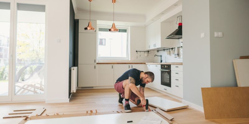 A person assembles furniture in a bright kitchen. They kneel on the floor with tools and white cabinet pieces. A person assembles furniture in a bright kitchen. They kneel on the floor with tools and white cabinet pieces.