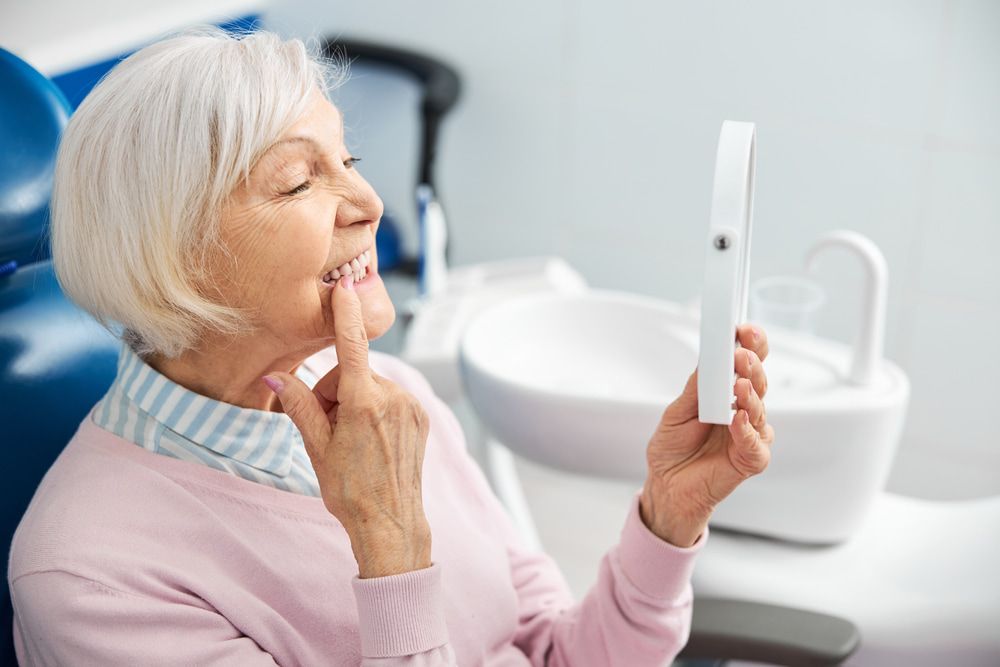 An Elderly Woman Is Sitting In A Dental Chair Looking At Her Teeth In A Mirror — Miami Denture Clinic In Burleigh Heads, QLD