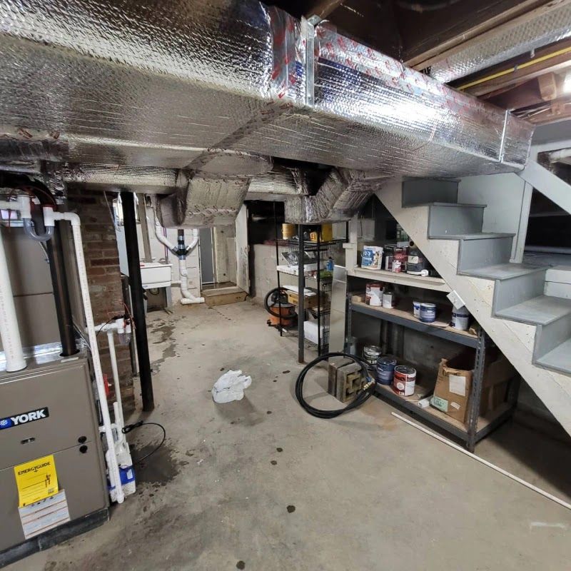 Basement with HVAC unit, ductwork, stairs, shelves with supplies, and a concrete floor.
