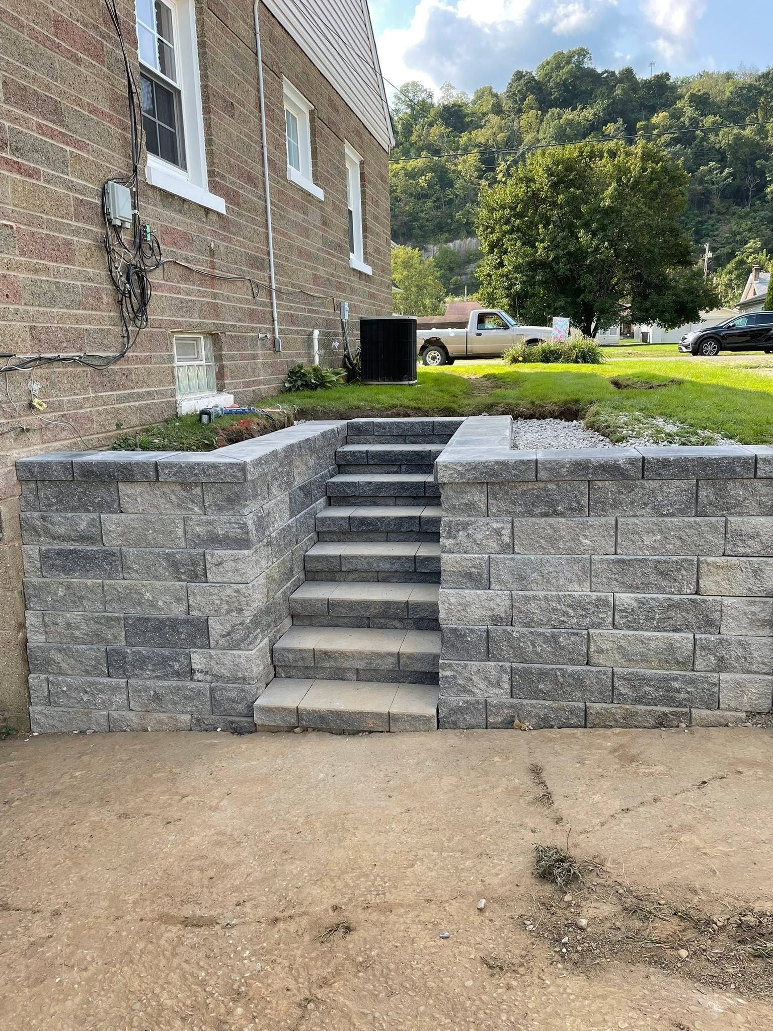 Stone steps and retaining wall leading up to a brick building, with a grassy yard and parked truck nearby.