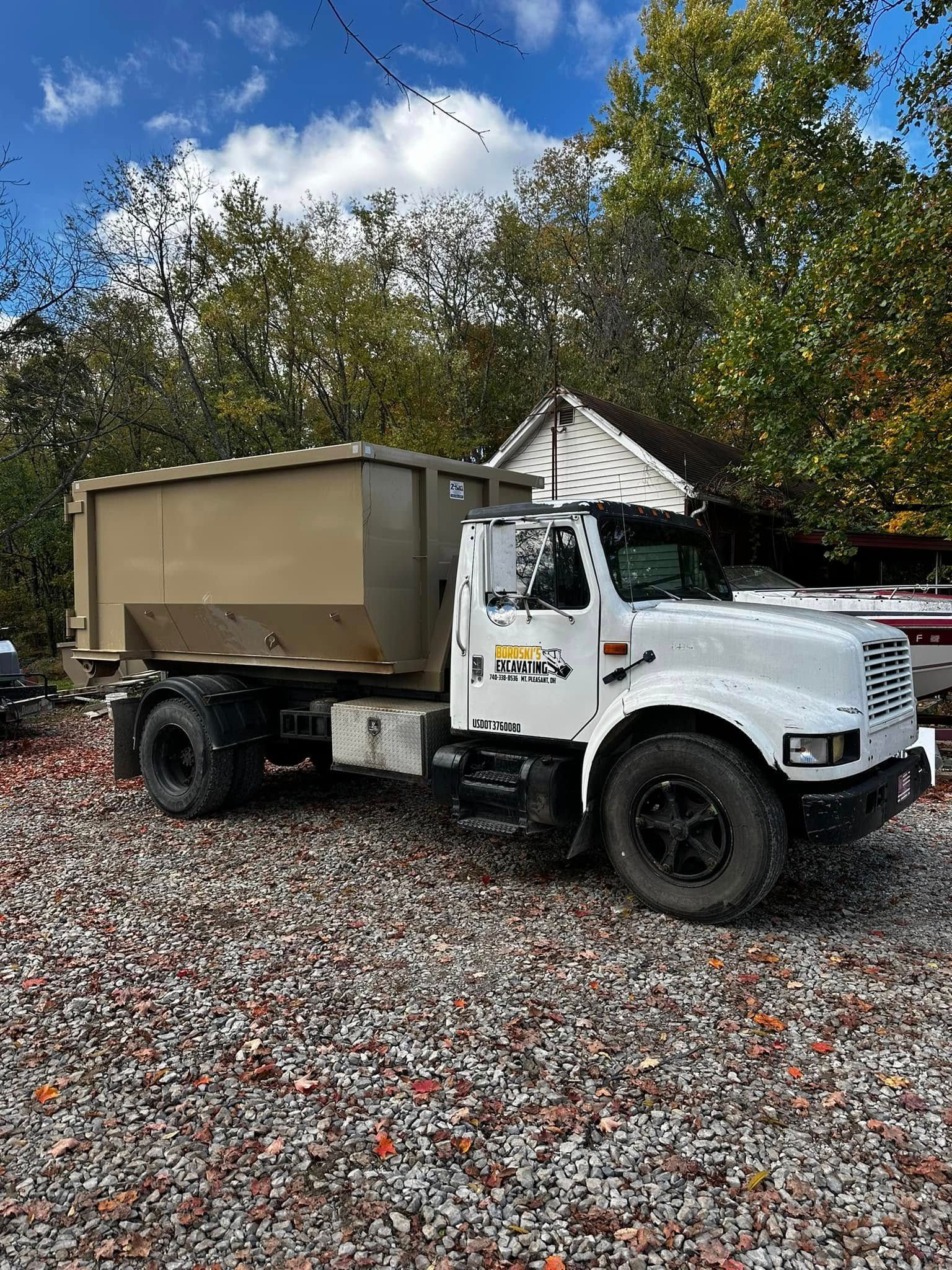 White dump truck with tan bed parked on gravel in front of a house and trees