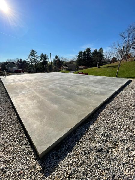 Newly poured concrete slab in a sunny outdoor lot with gravel edges and grassy hillside nearby