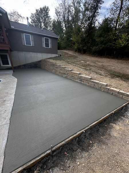 Freshly poured concrete patio beside a house, bordered by stone edging and gravel yard