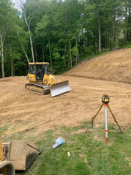 Bulldozer grading a dirt hillside on a wooded construction site, with surveying tripod in foreground