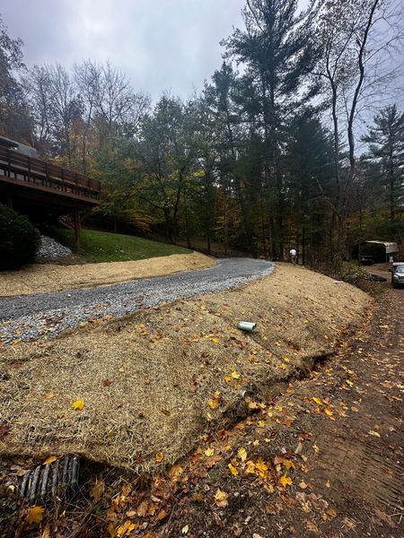 Gravel driveway beside a wooded hillside under overcast skies, with a bridge on the left.