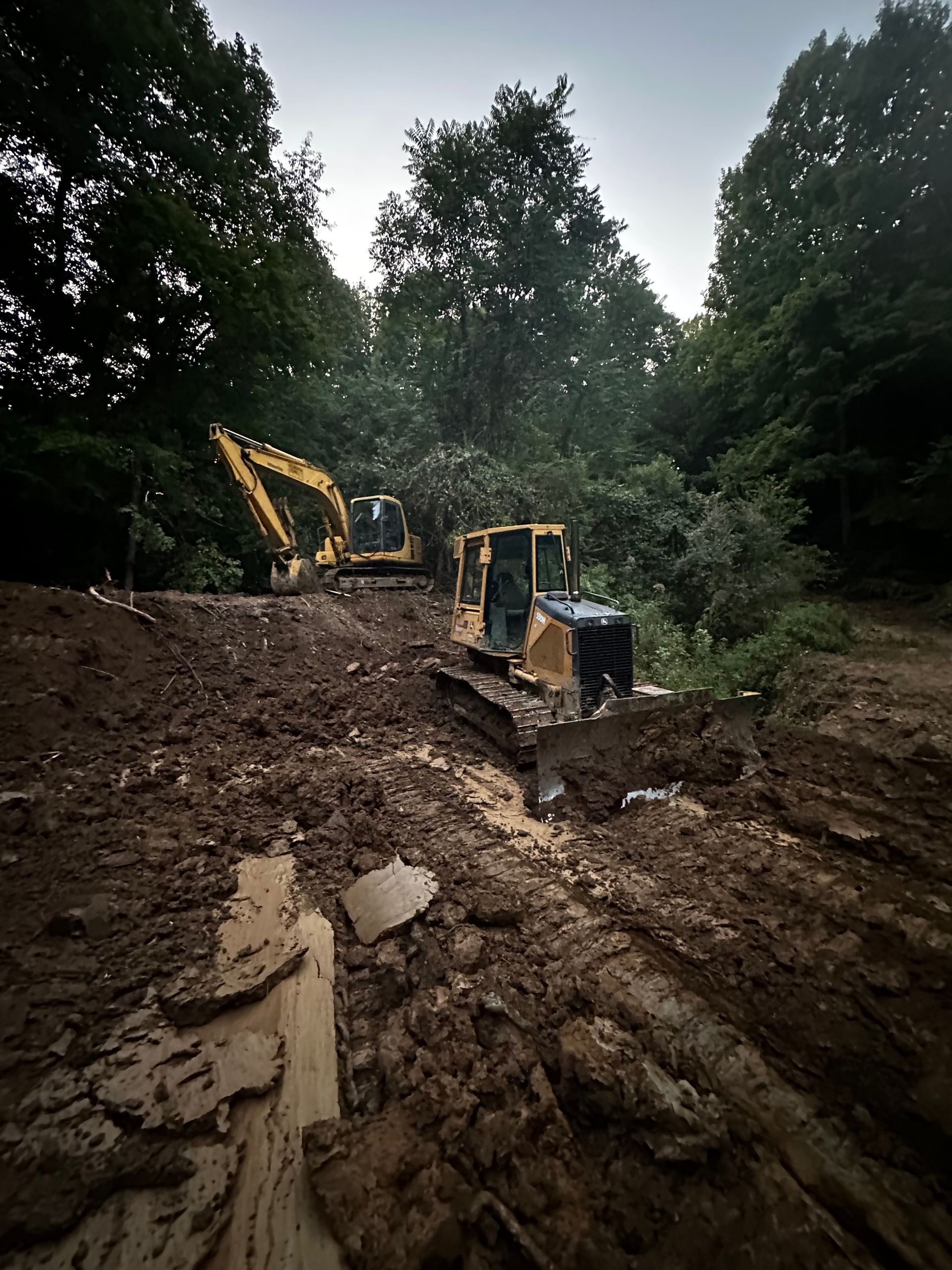 Two excavators working in a muddy dirt clearing surrounded by trees