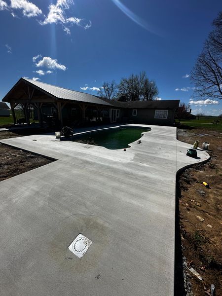 Concrete patio with a dark-green pool beside a covered house under a bright blue sky.