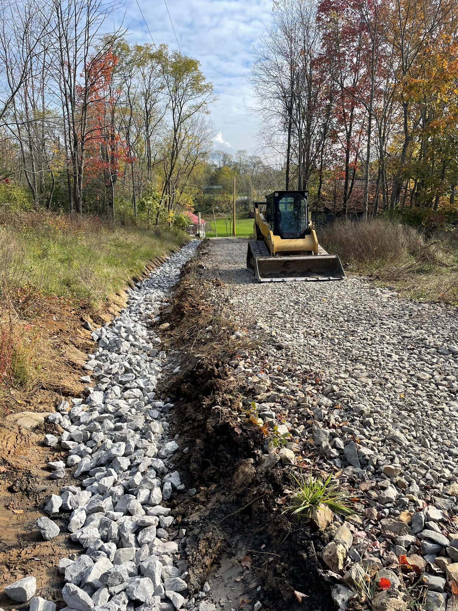 Small excavator on a gravel road beside a rock-lined ditch in a wooded area