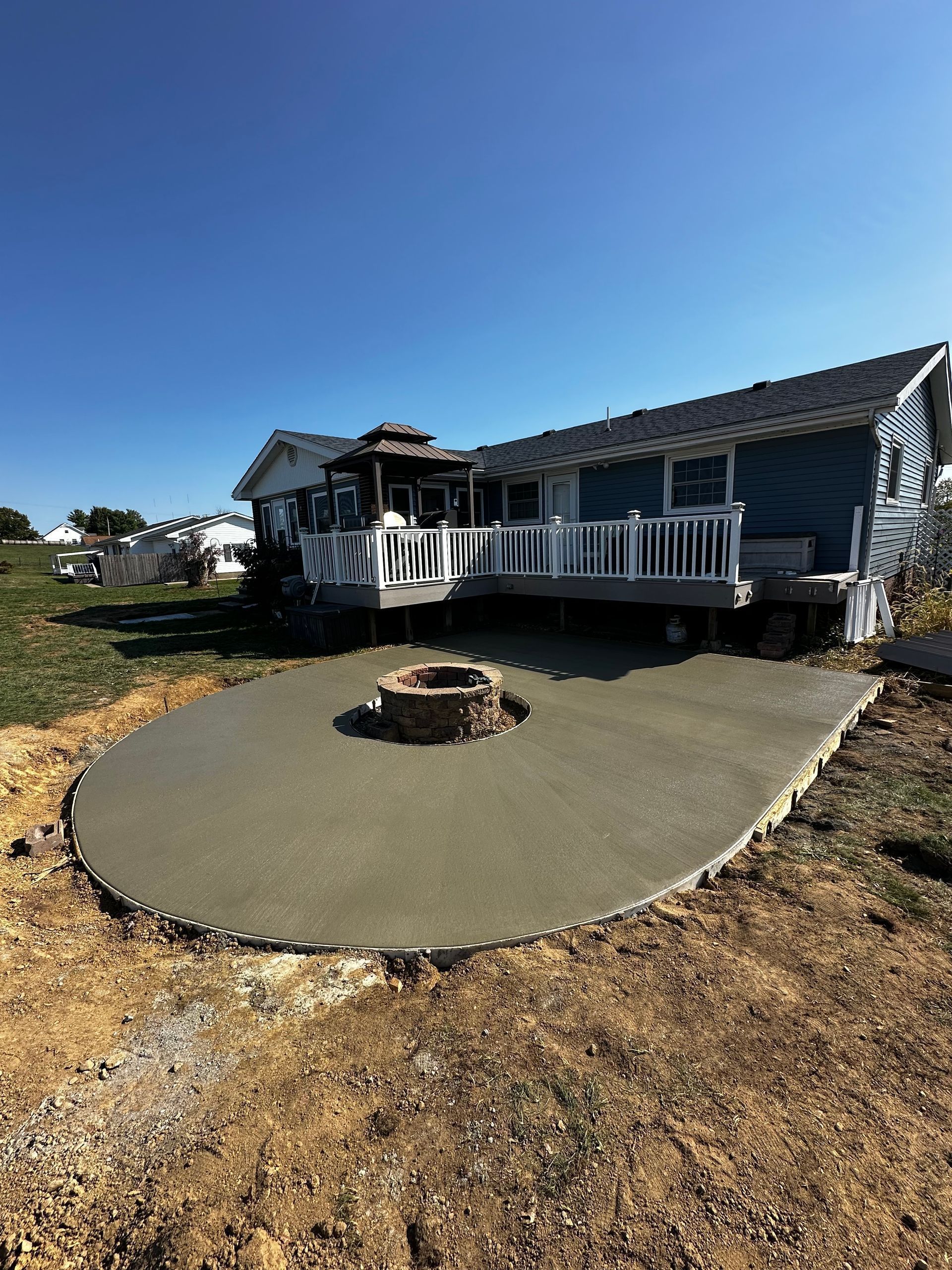Single-story house with a white deck and a large circular concrete patio with a fire pit in the yard.