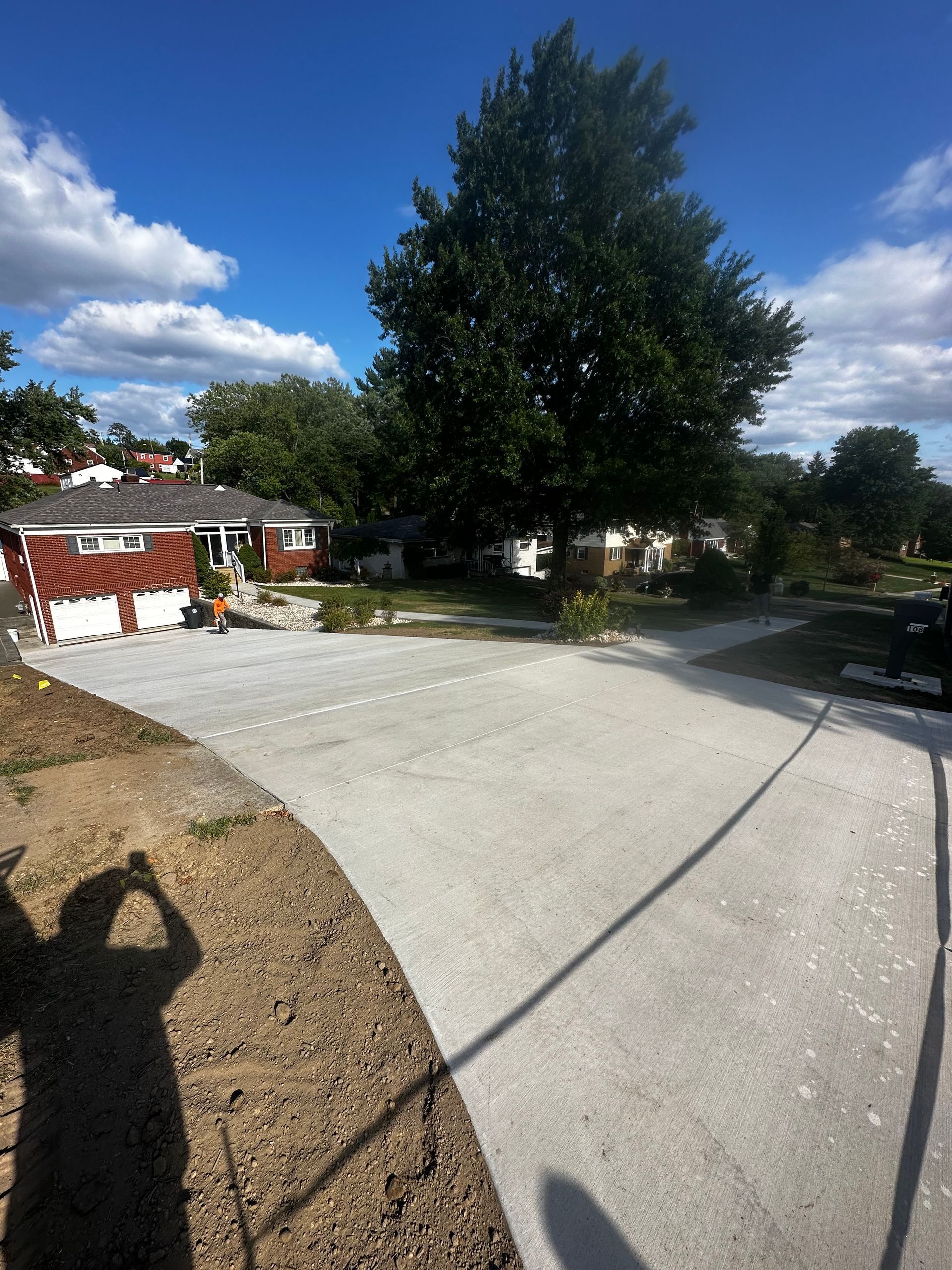 Large concrete driveway with a tall tree, houses, and blue sky with clouds
