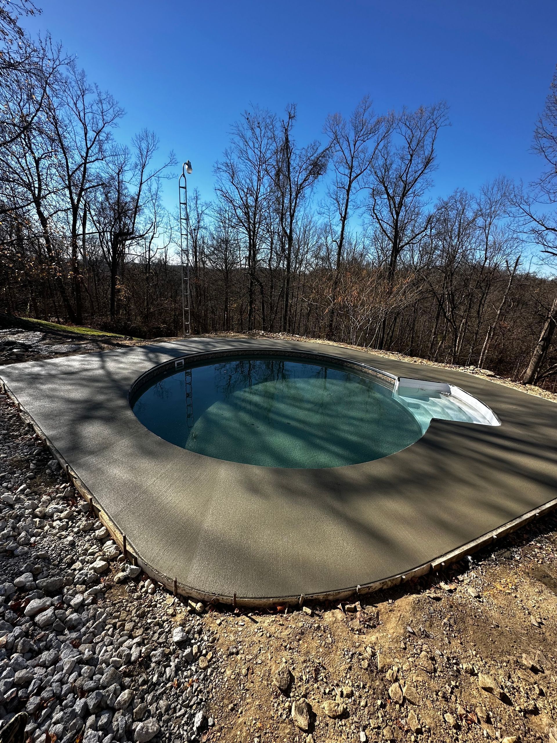 Small outdoor hot tub with blue water beside a gravel path, surrounded by leafless trees under a clear sky