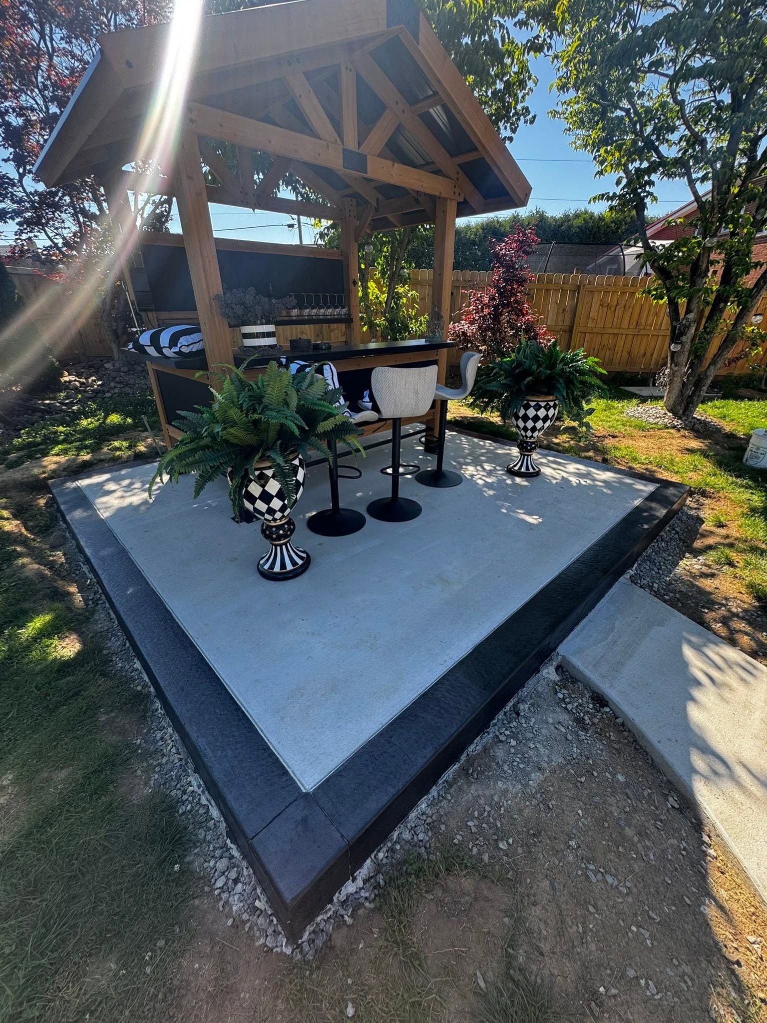 Outdoor gazebo with a small raised concrete platform, potted plants, and a decorated tree in a sunny garden
