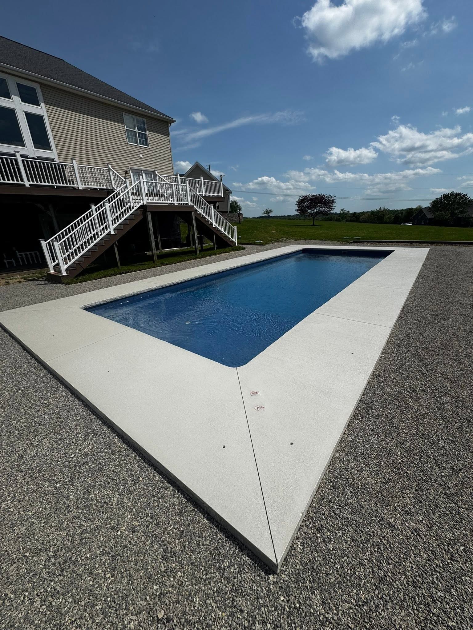 Backyard pool beside a house with white deck and gravel, under a blue sky