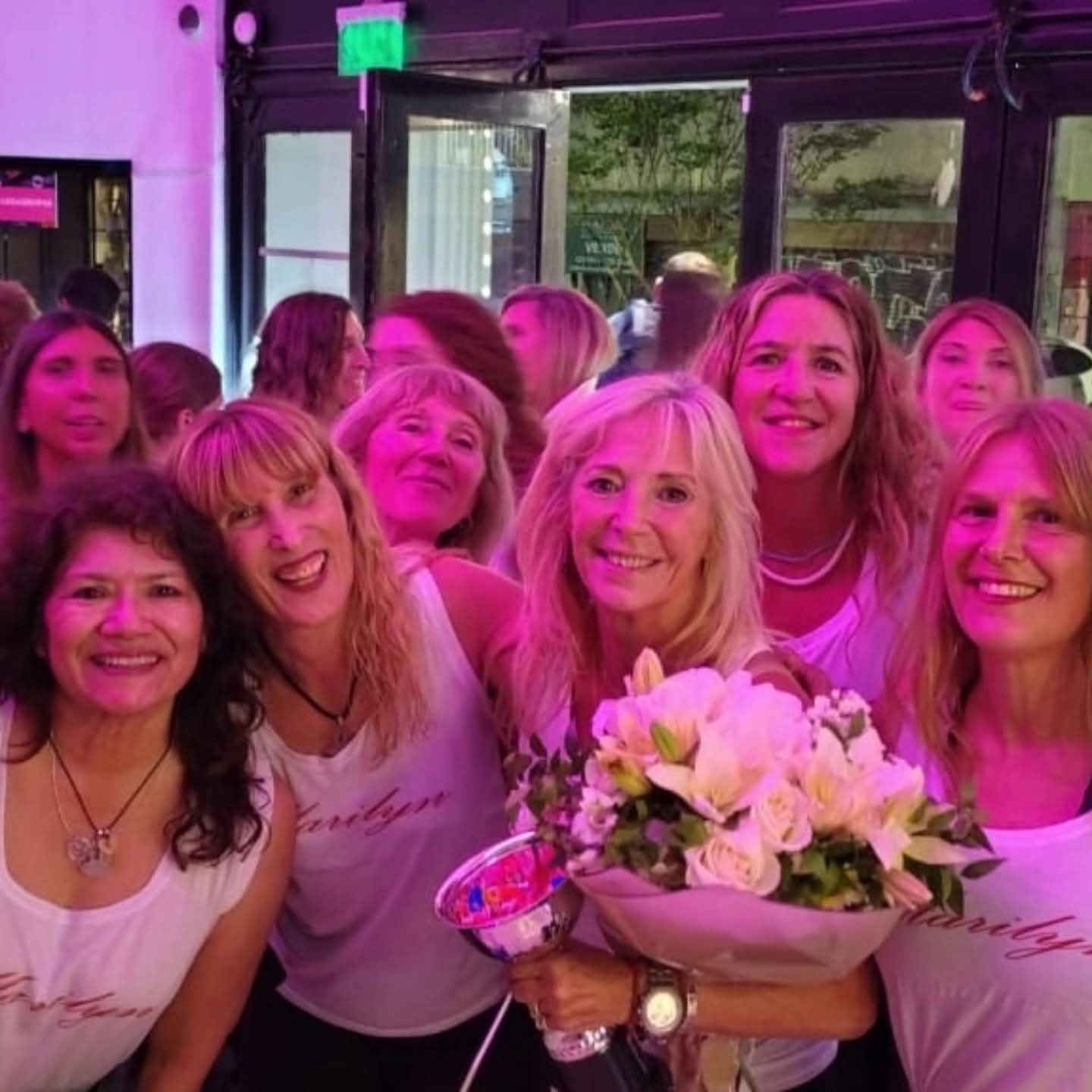 Grupo de mujeres sonrientes con camisas blancas y flores en un espacio interior iluminado con luz rosa