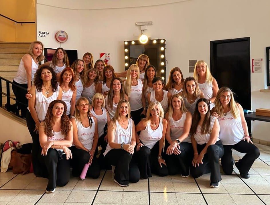 Grupo de mujeres posando con camisetas blancas a juego y pantalones negros en un salón.