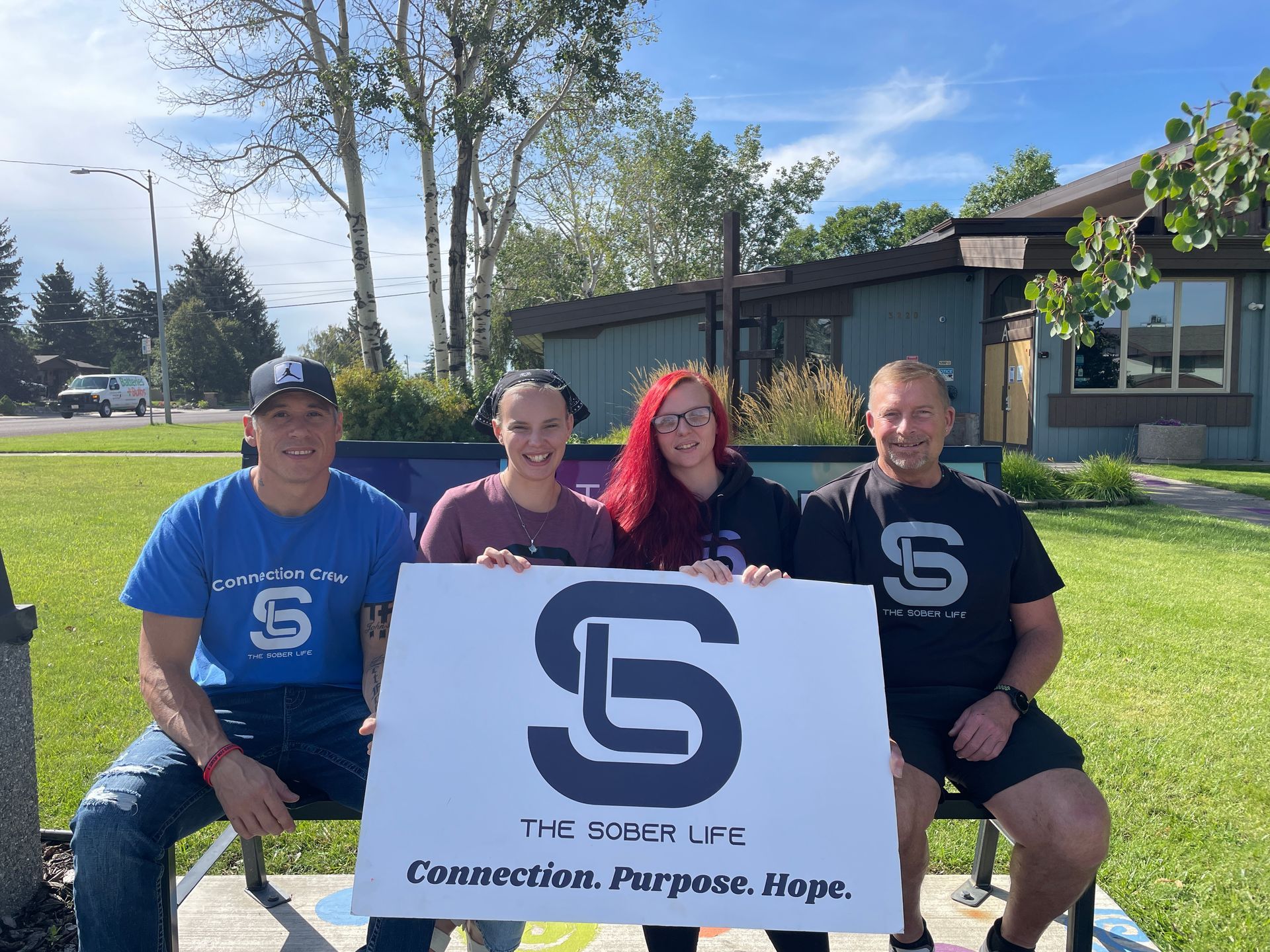 Four people sit on a park bench, holding a sign that reads 