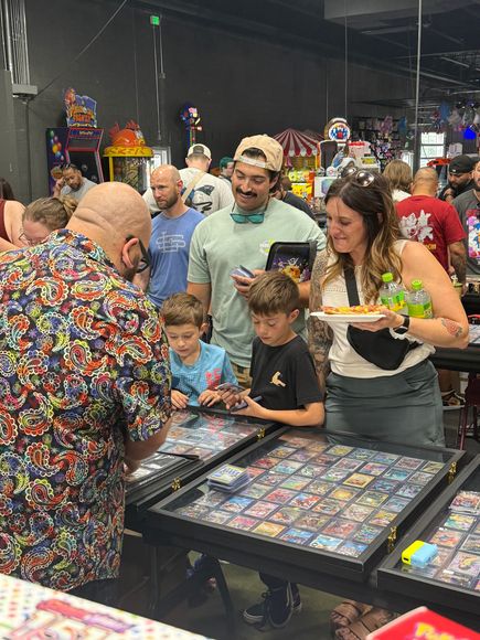 People browsing trading cards at a convention, including children.  A man in a colorful shirt is showing something to them.