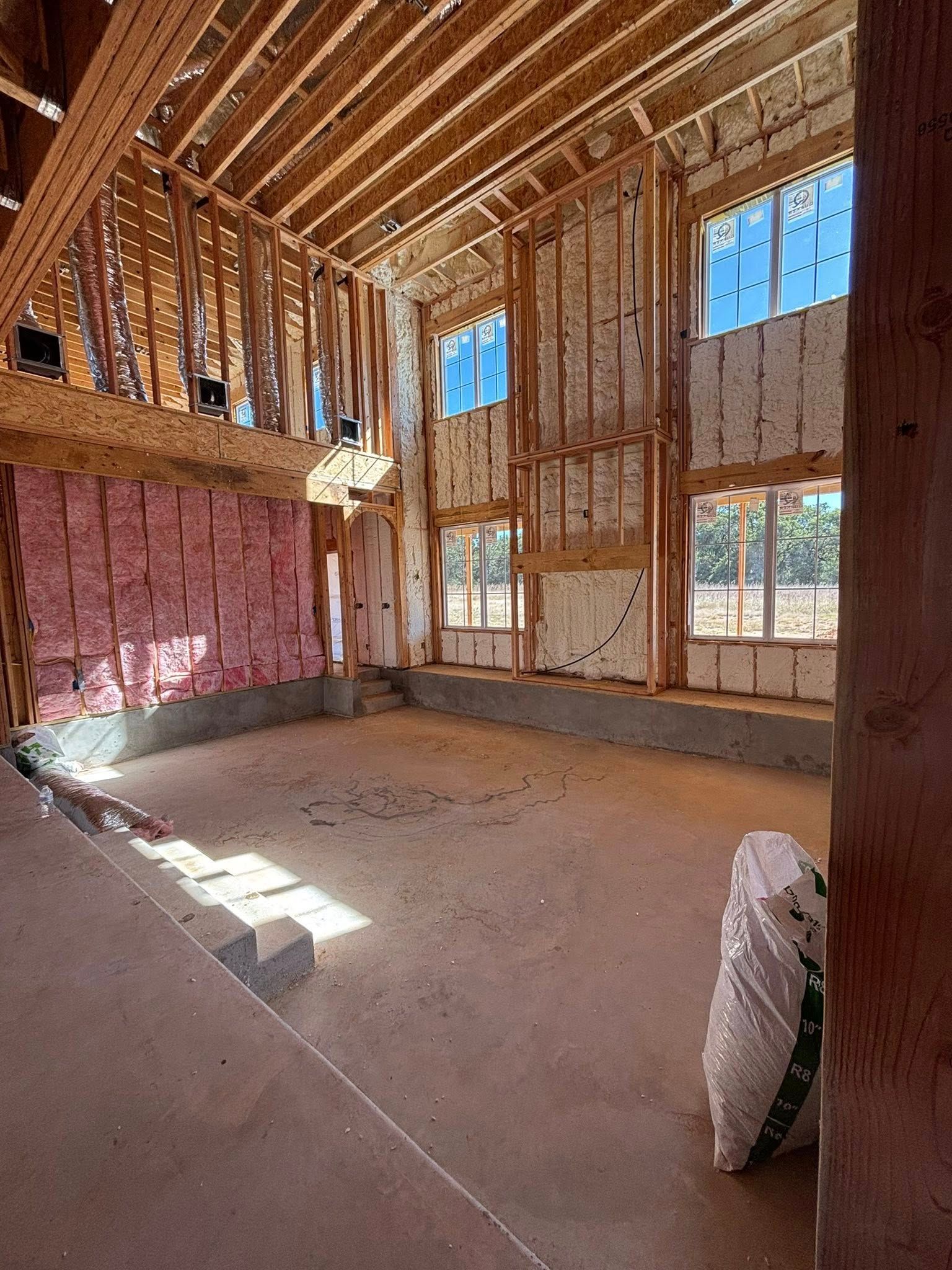 Interior of a house under construction with exposed wood framing, pink insulation, and sunlight through windows.