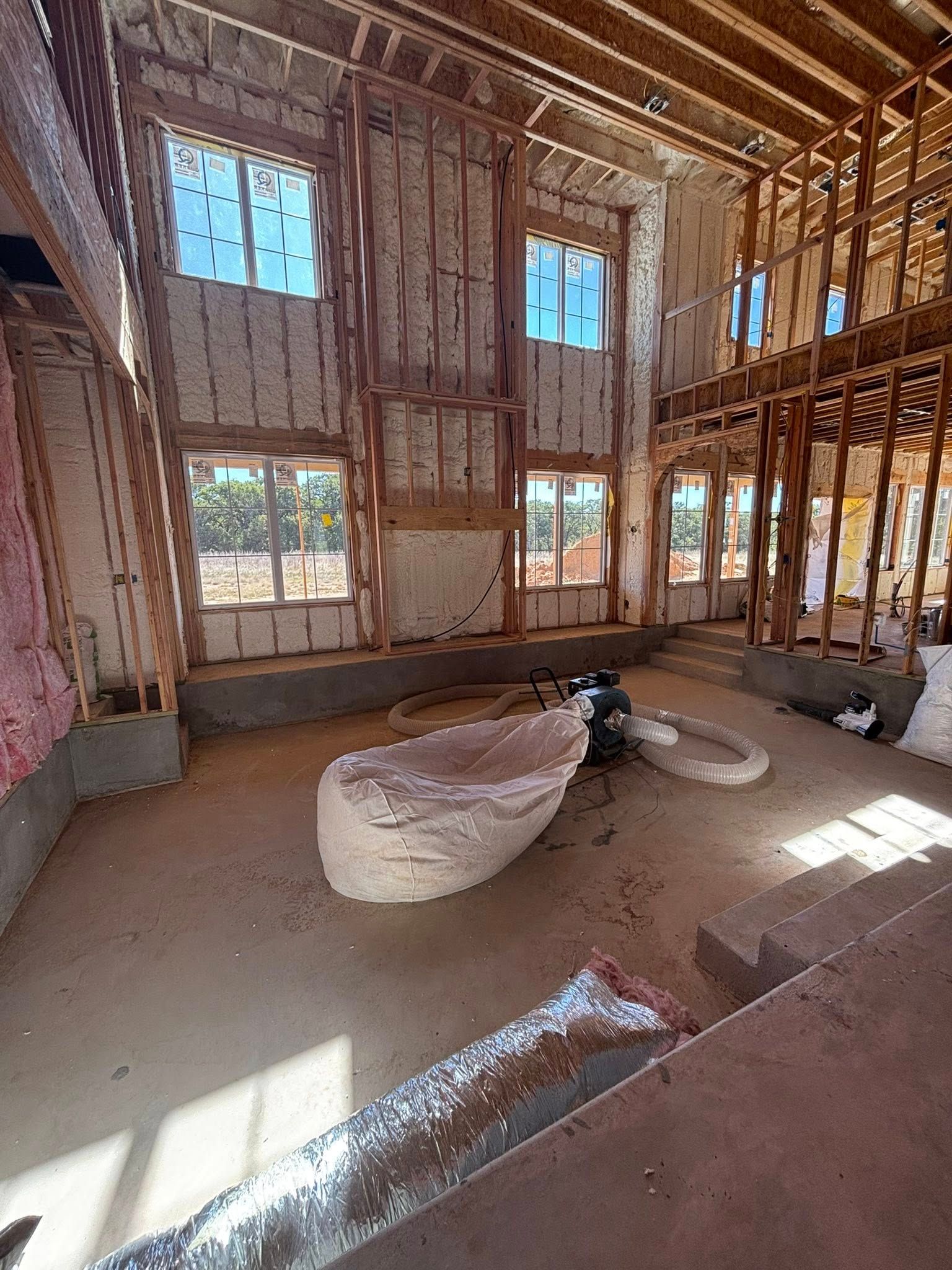 Interior of a framed, unfinished room with exposed studs, windows, and a bag of materials on the floor
