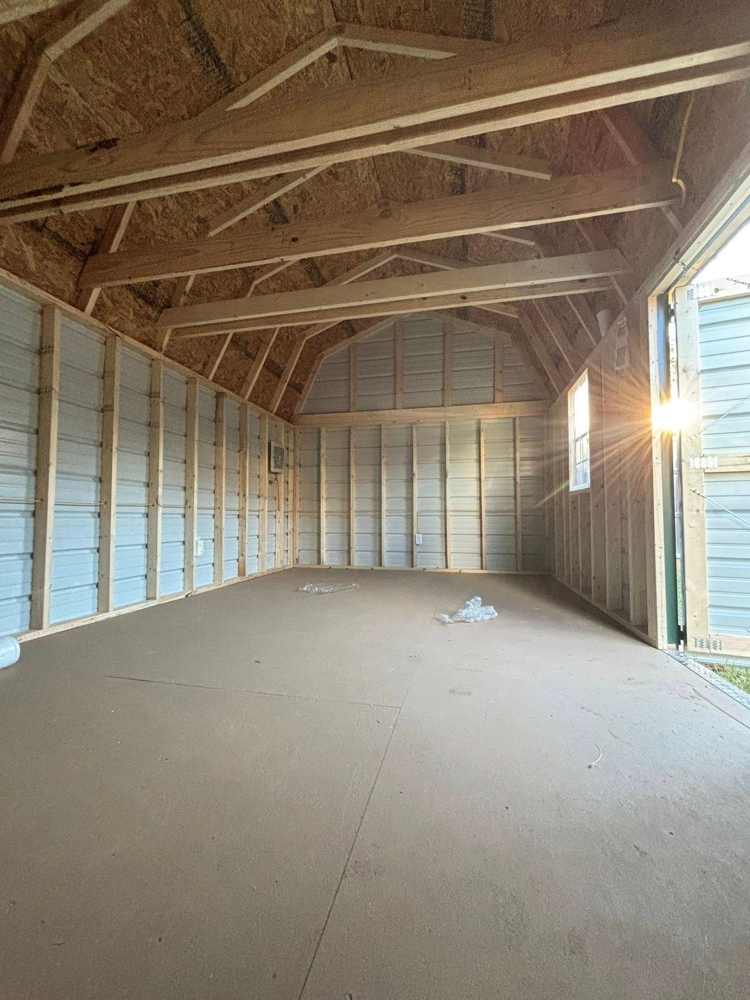 Empty unfinished shed interior with exposed wooden rafters and sunlight from the open doorway.