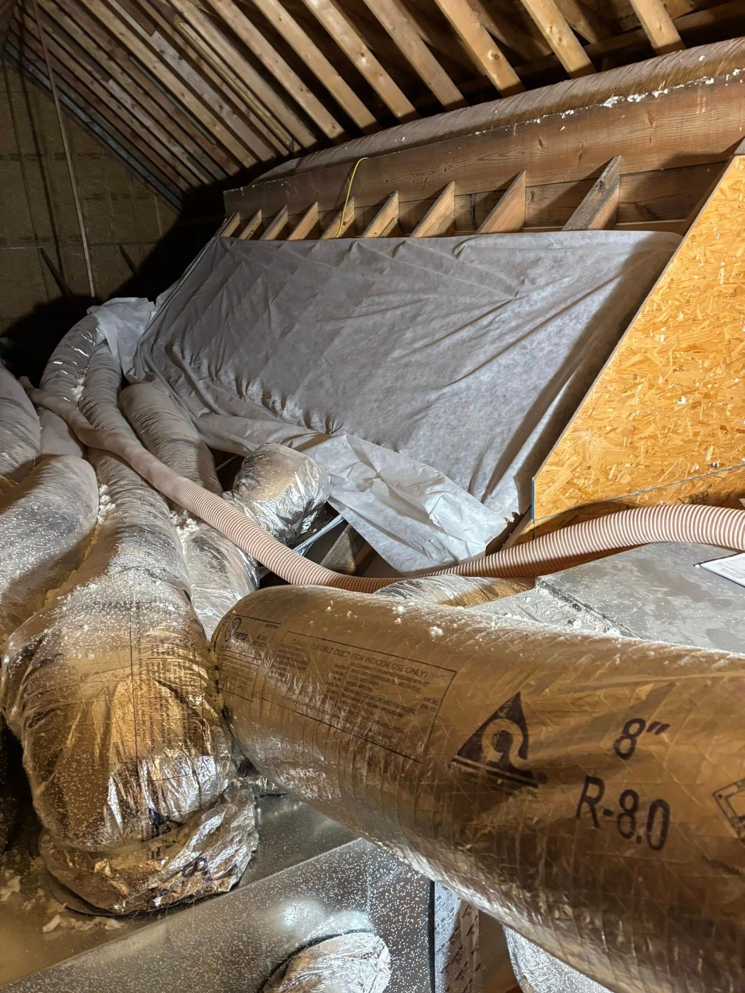 Insulated attic with exposed beams, ducting, and covered equipment amid scattered insulation debris.