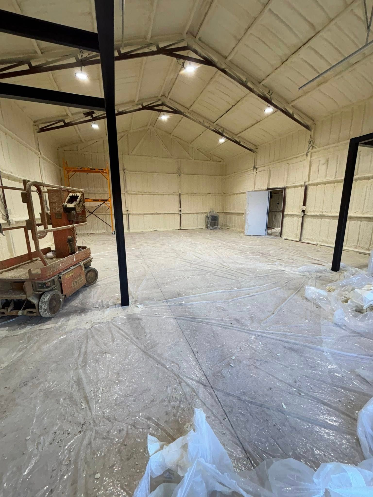 Empty warehouse interior with plastic-covered floor, exposed beams, and construction equipment on the left.