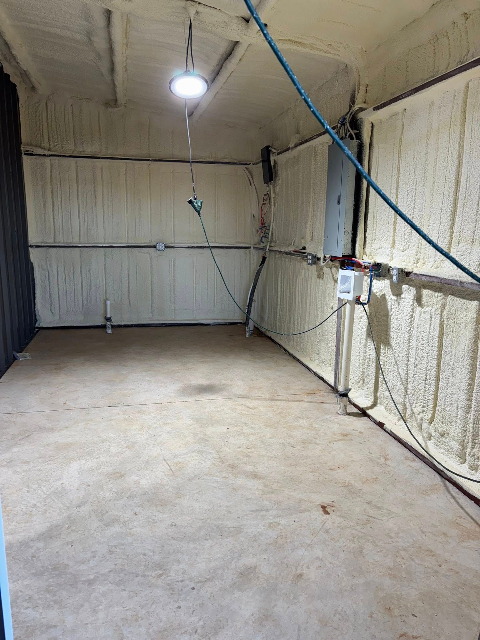 Empty unfinished room with concrete floor, white insulated walls, exposed wiring, and a hanging light bulb.