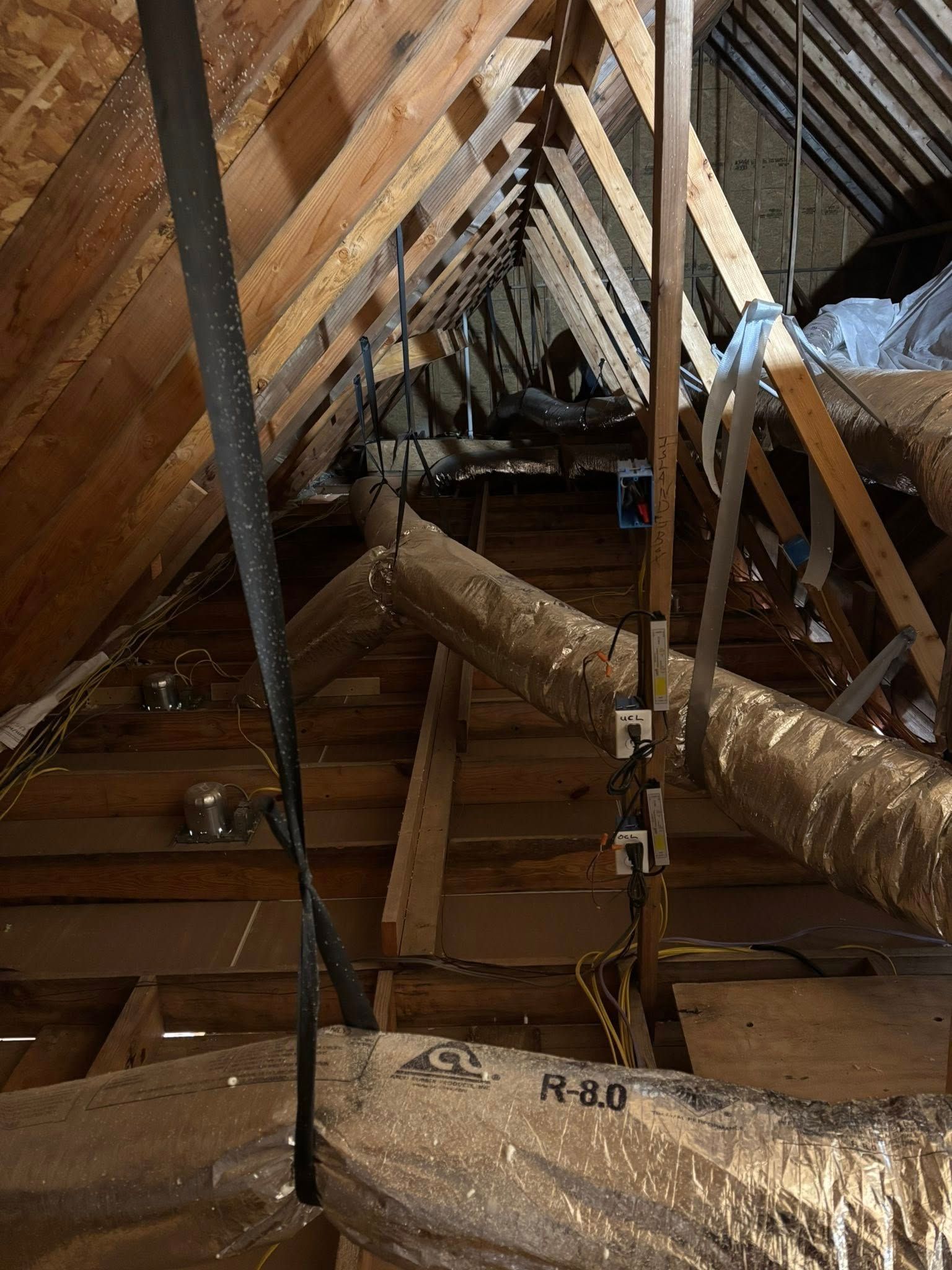 Attic with exposed wooden beams, insulation, and hanging black pipes