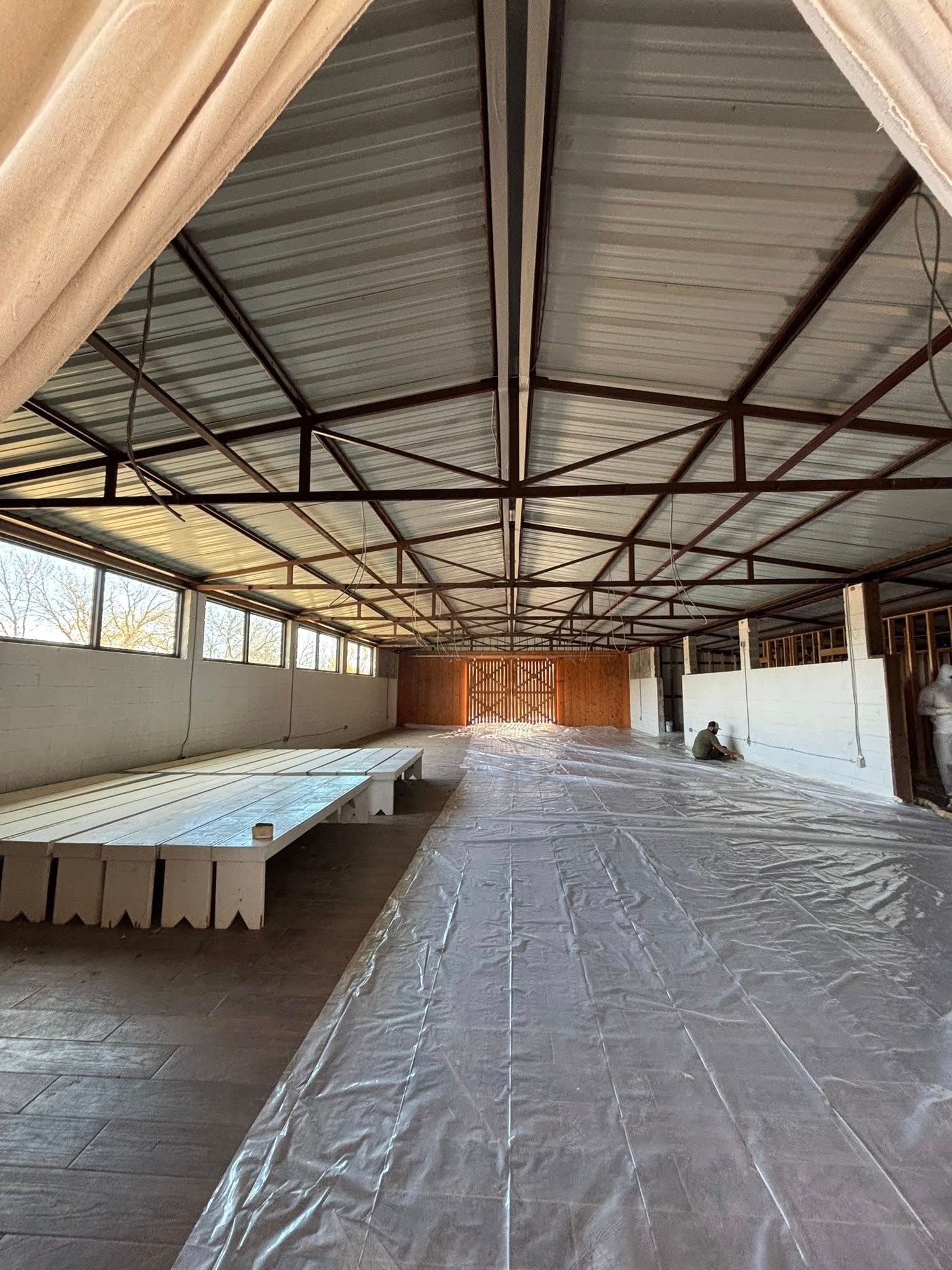 Empty indoor hall with long tables, plastic-covered floor, and corrugated metal roof.