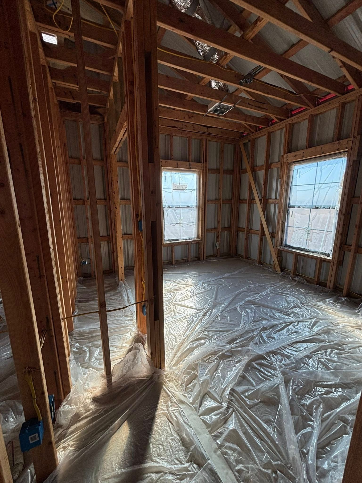 Interior of a framed house under construction, with exposed wood studs, plastic-covered floor, and two windows.