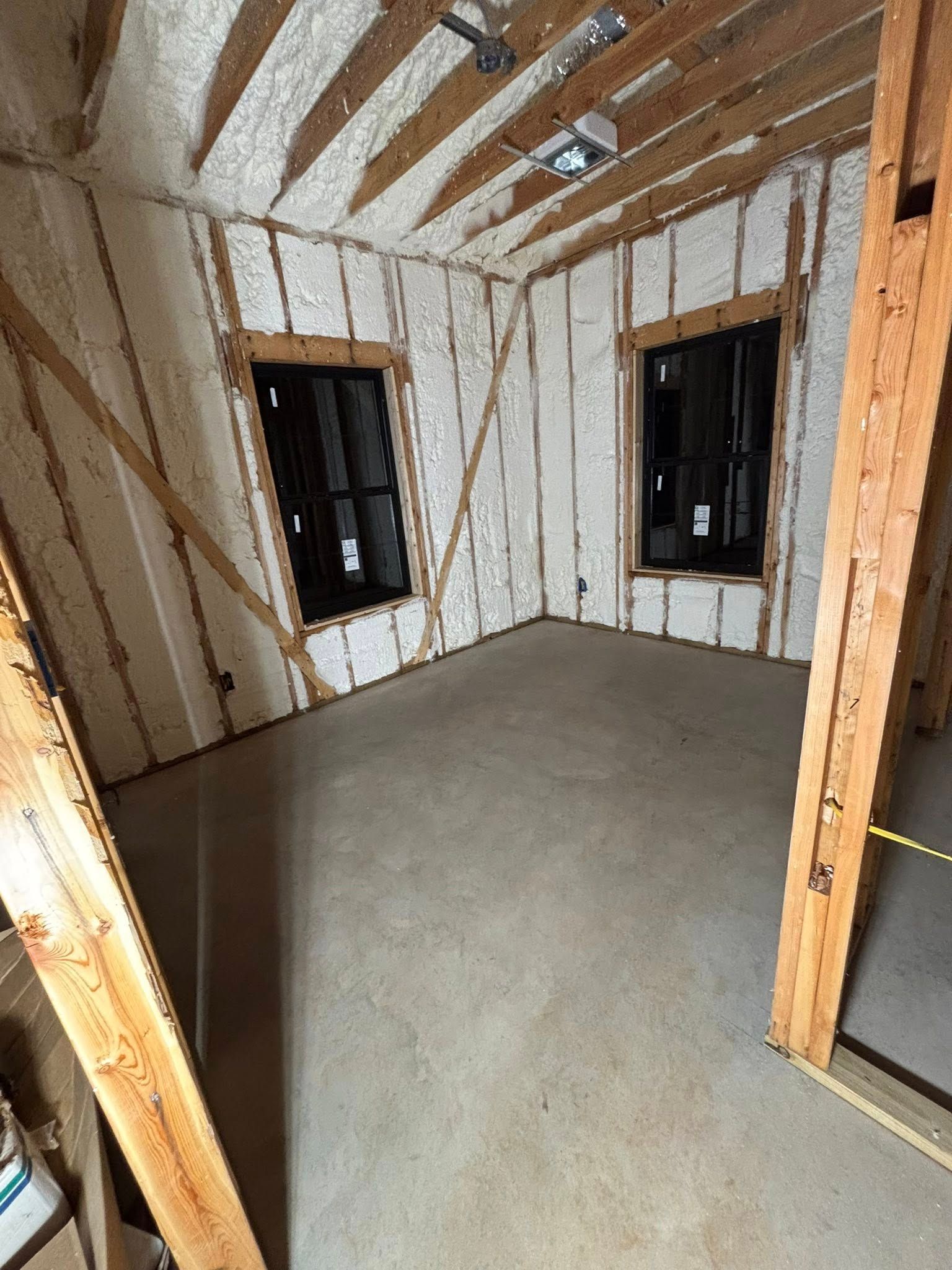 Empty unfinished room with exposed framing, insulation, dark windows, and a bare concrete floor.
