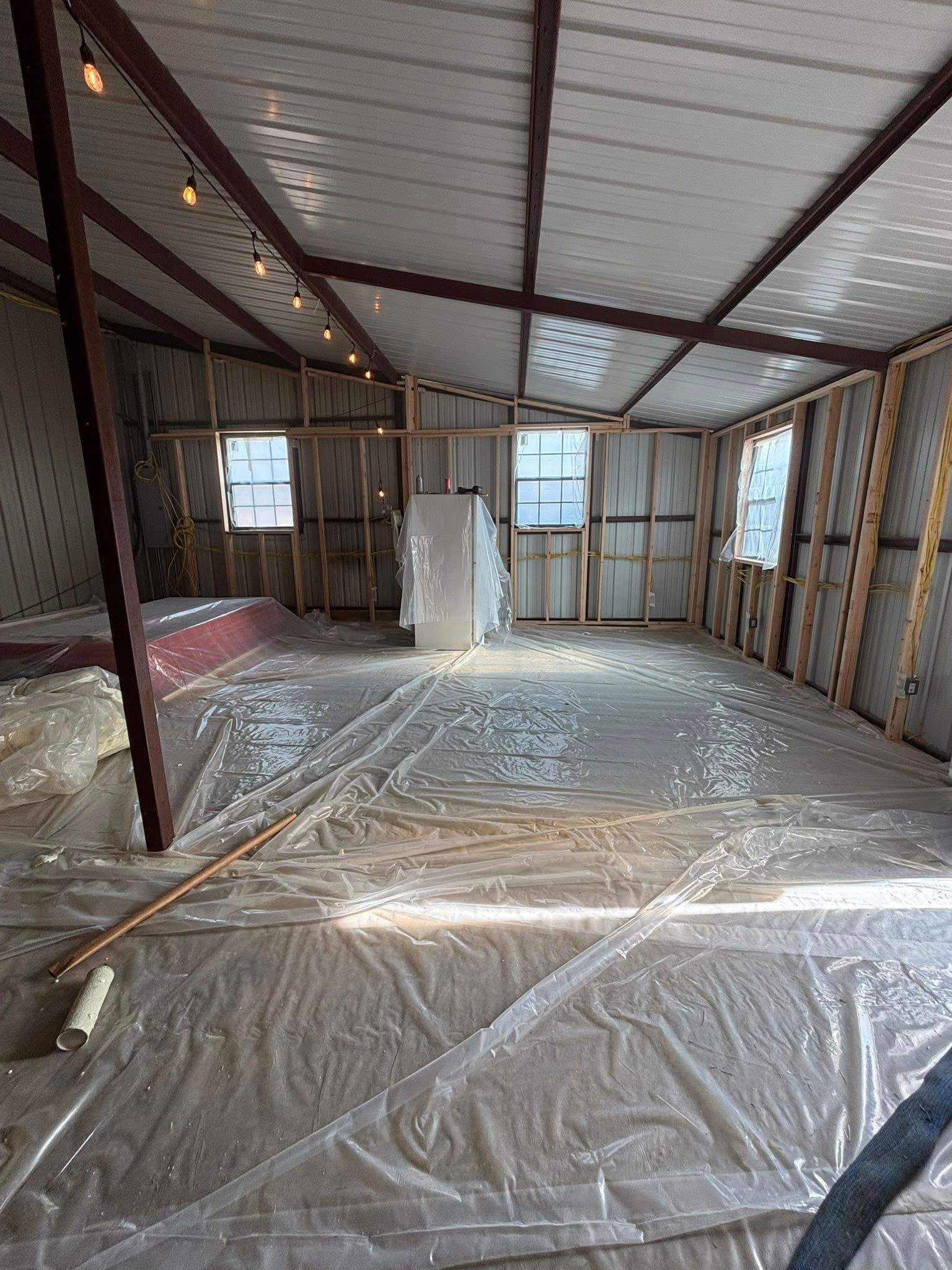 Partially renovated attic room with exposed framing, plastic-covered floor, and windows letting in daylight