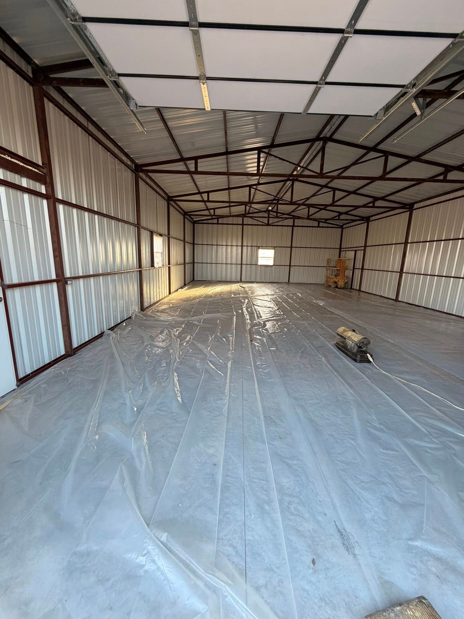 Empty metal workshop with plastic-covered floor, exposed beams, and sunlight streaming through side windows