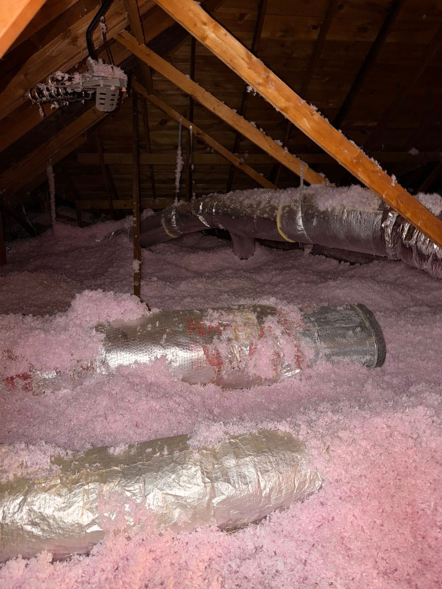 Insulated attic with exposed wooden beams and metal ducts above pink insulation batts