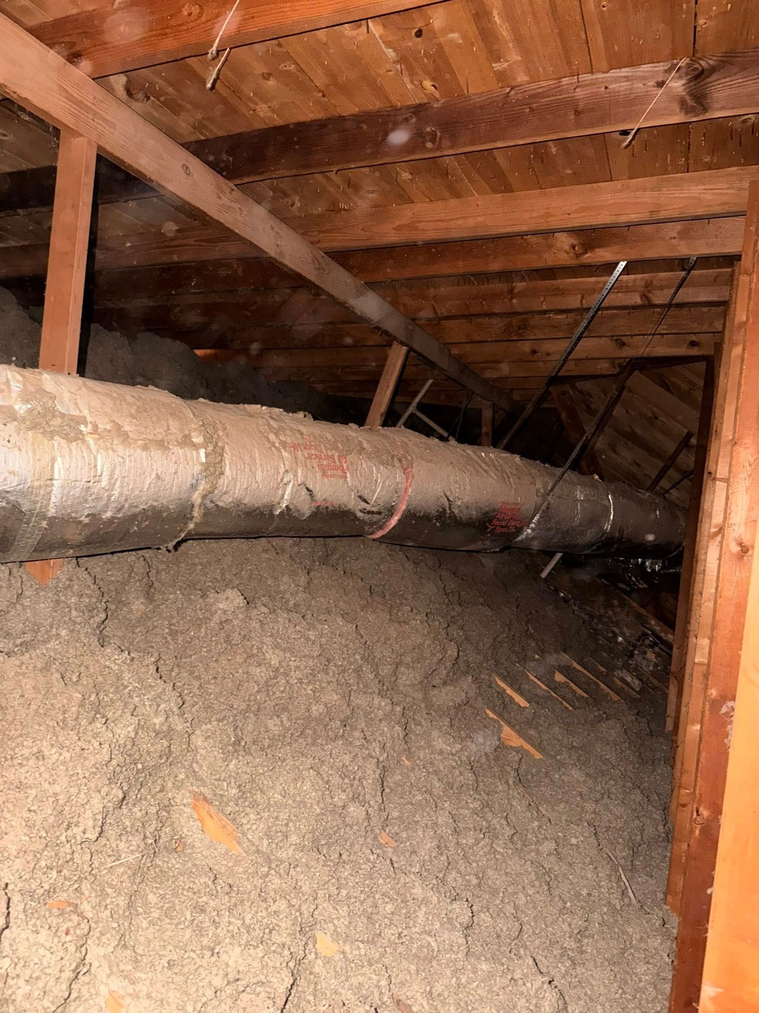 Inside attic crawlspace with exposed wooden beams and a large horizontal pipe over insulation debris.